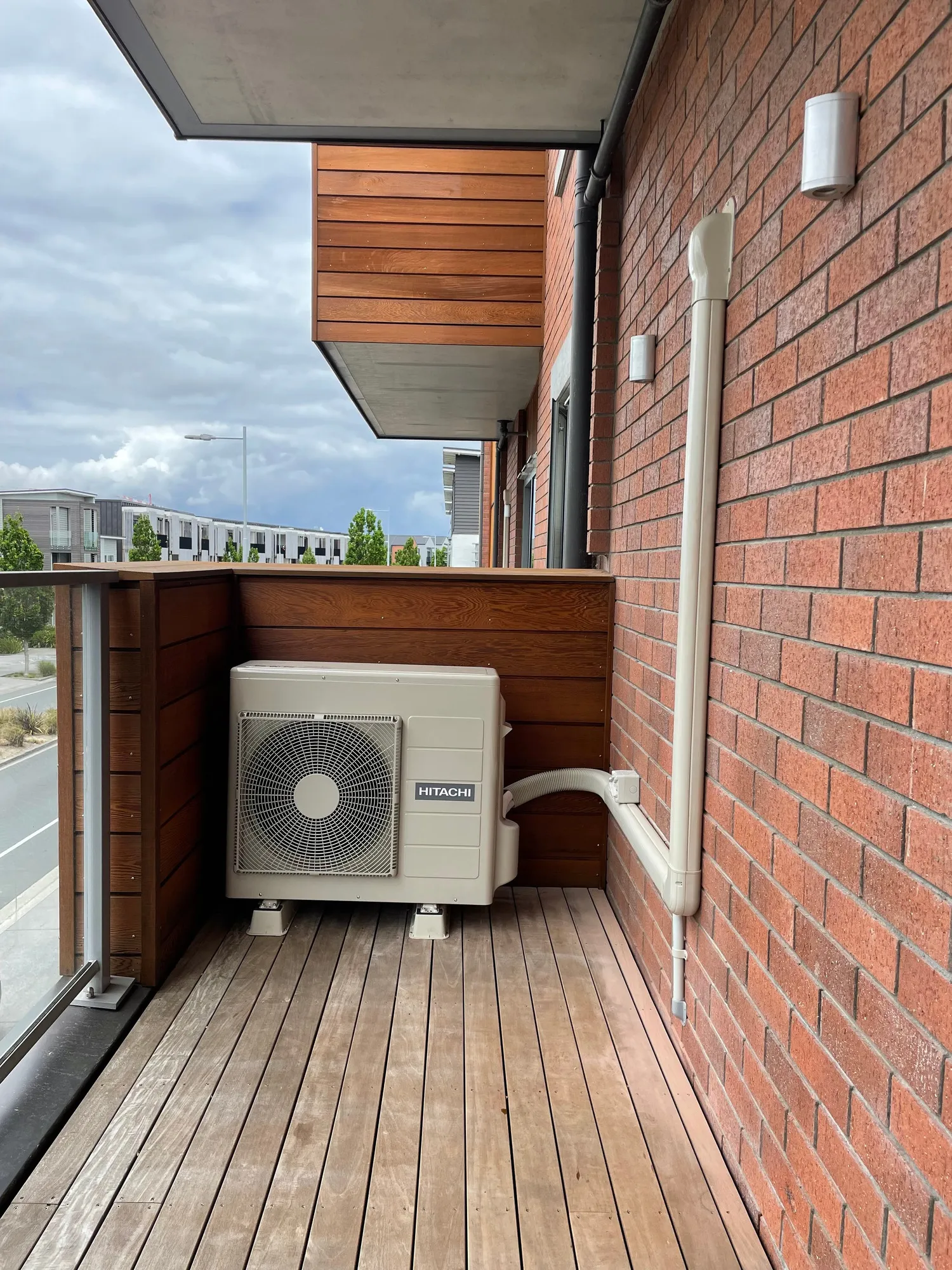 A heat pump outdoor unit on a timber balcony, with a white pipe cover running up a red brick wall.