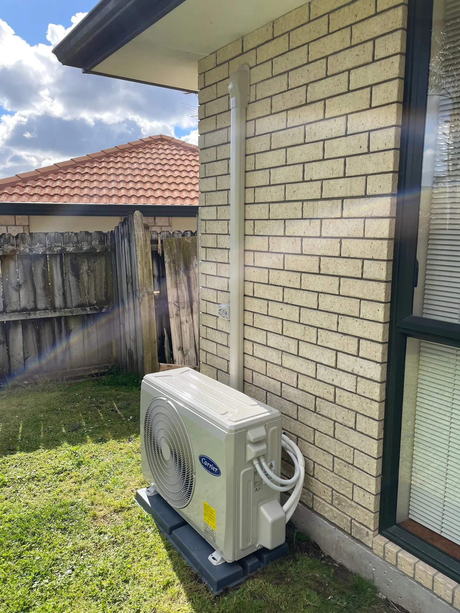 A heat pump outdoor unit on a base on the lawn beside a cream brick wall, with a white pipe cover running up to the eaves.