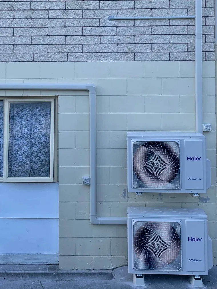 Two heat pump outdoor units, one above the other, on a painted block wall with white pipe covers.
